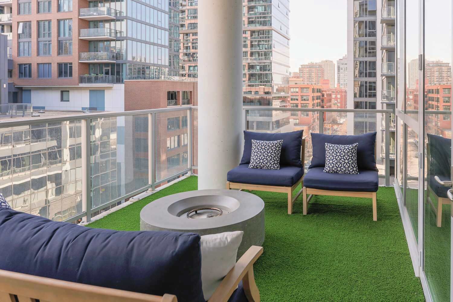 Een balkon met twee loungestoelen, een ronde tafel en een kleine vuurkuil. Het balkon heeft een glazen balustrade en uitzicht op omliggende gebouwen.