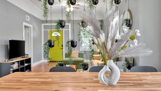 A modern dining area with a wooden table, white vase with dried plants, hanging plants, and a TV in the background. - The Green Door in Ybor  (Tampa)
