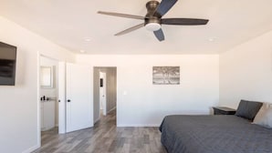 Une chambre à coucher avec un lit, un ventilateur de plafond, un tableau au mur et un couloir menant à une salle de bain.