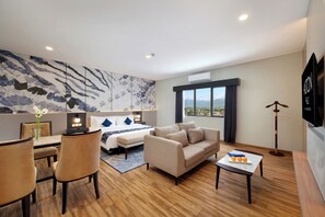 Habitación de hotel moderna con cama, sofá, zona de comedor y un amplio ventanal con vistas a la ciudad.