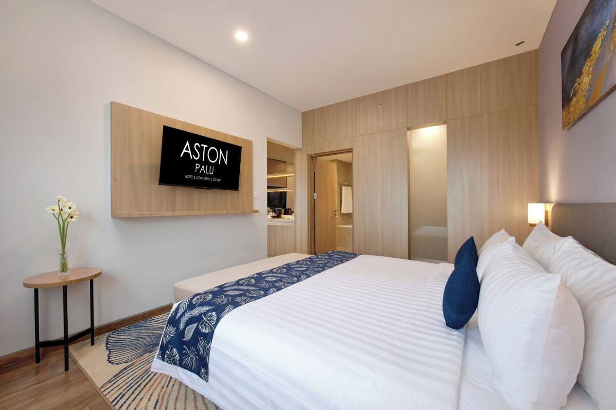Une chambre d’hôtel avec un grand lit, un téléviseur fixé au mur, une petite table avec un vase et une salle de bain visible à travers une porte coulissante.