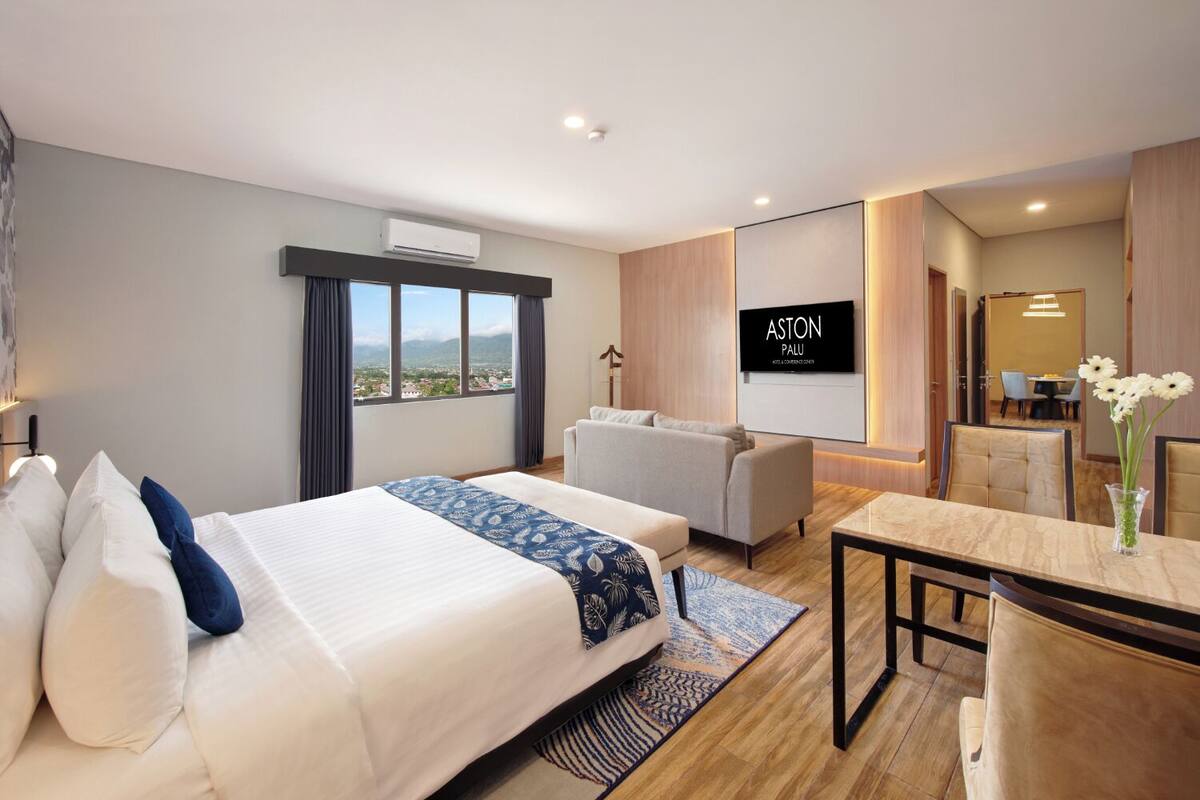 Une chambre d’hôtel moderne dotée d’un grand lit, d’un canapé, d’une table à manger et d’un téléviseur affichant le logo « ASTON PALLU ».