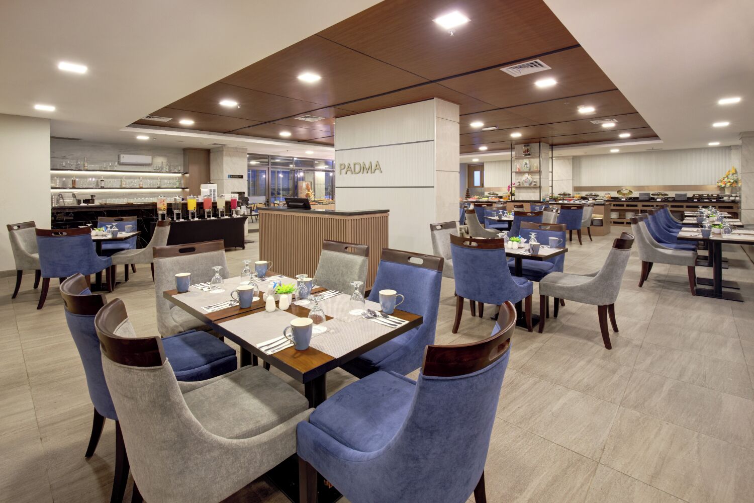 A modern dining area with multiple tables and chairs, a reception desk, and a kitchen area in the background.