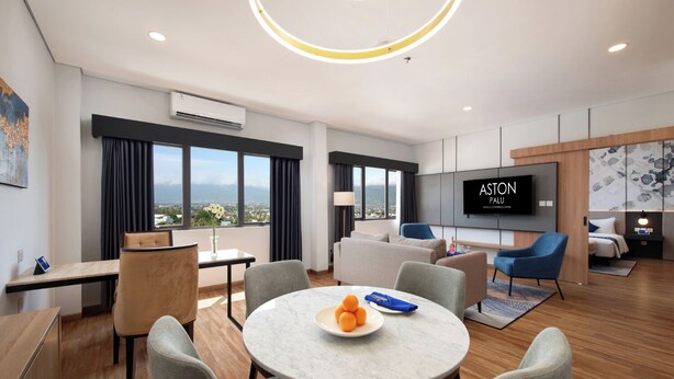 A modern hotel room with a dining area, a living area with a TV, and a view of the city. - Aston Palu Hotel & Conference Center (Palu)