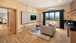 A modern hotel room with a flat-screen TV, a sofa, a small coffee table, and a view of the city through large windows. - Aston Palu Hotel & Conference Center (Palu)