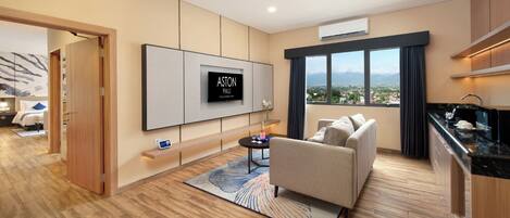 A modern hotel room with a flat-screen TV, a sofa, a small coffee table, and a view of the city through large windows.