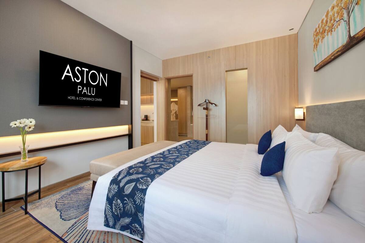 Une chambre d’hôtel avec un lit, une table de chevet ornée d’un vase de fleurs, un téléviseur à écran plat fixé au mur et un tableau au-dessus du lit.