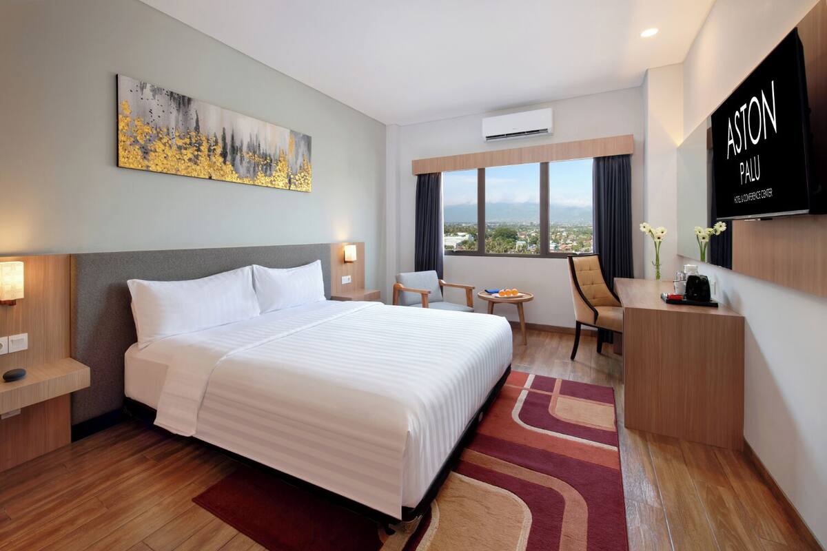 Une chambre d’hôtel avec un grand lit, un bureau, une chaise, une petite table avec des fruits et un téléviseur affichant le nom de l’hôtel.