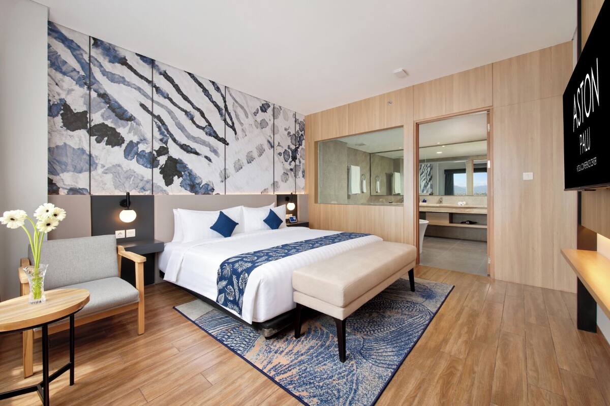 Une chambre d’hôtel moderne dotée d’un grand lit, d’un coin salon avec un canapé et des fauteuils, d’une petite table sur laquelle est posé un vase de fleurs, d’un grand miroir et d’un téléviseur fixé au mur.