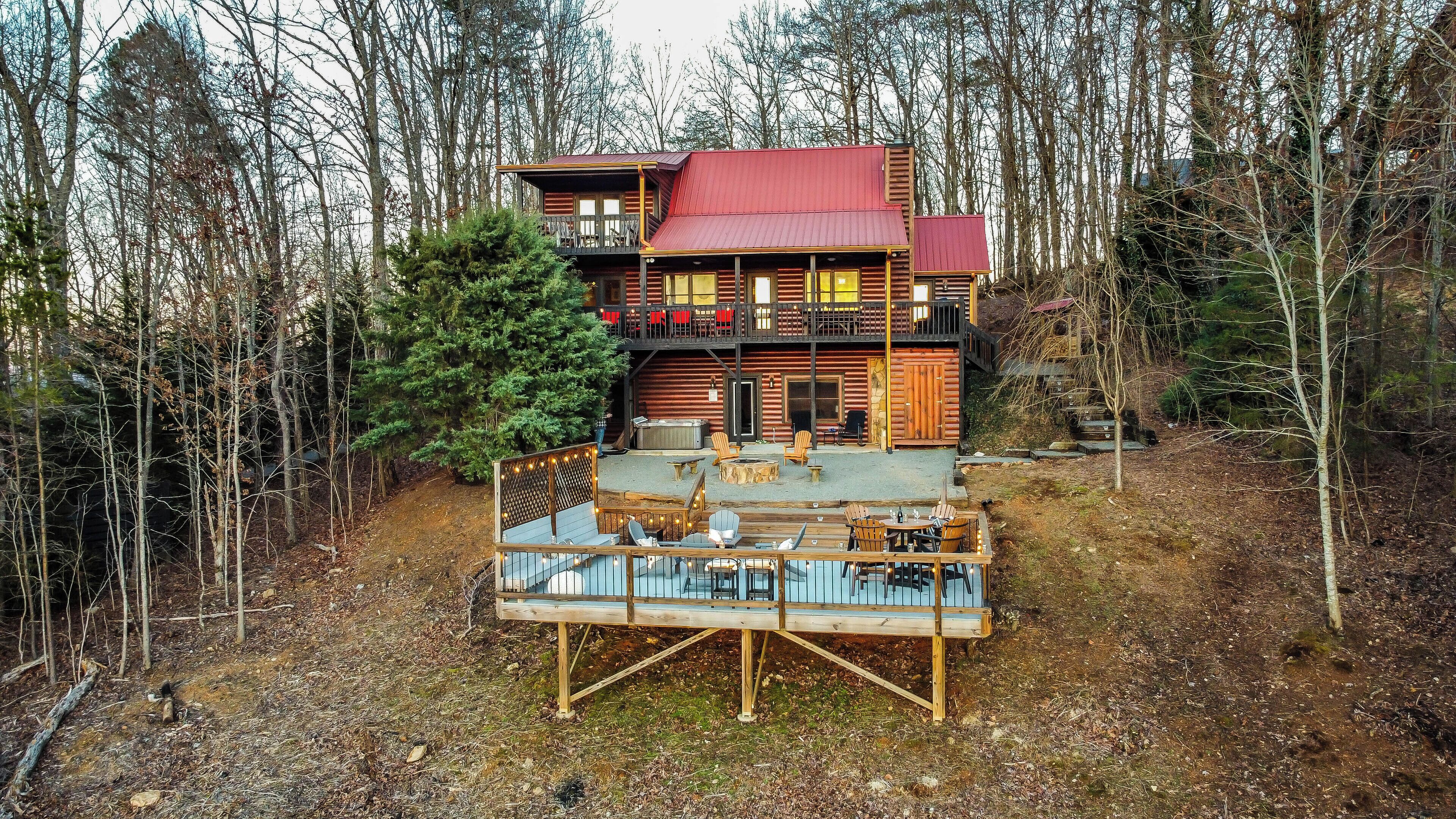 A log cabin with a red roof, a deck with outdoor seating, and a forested backdrop.