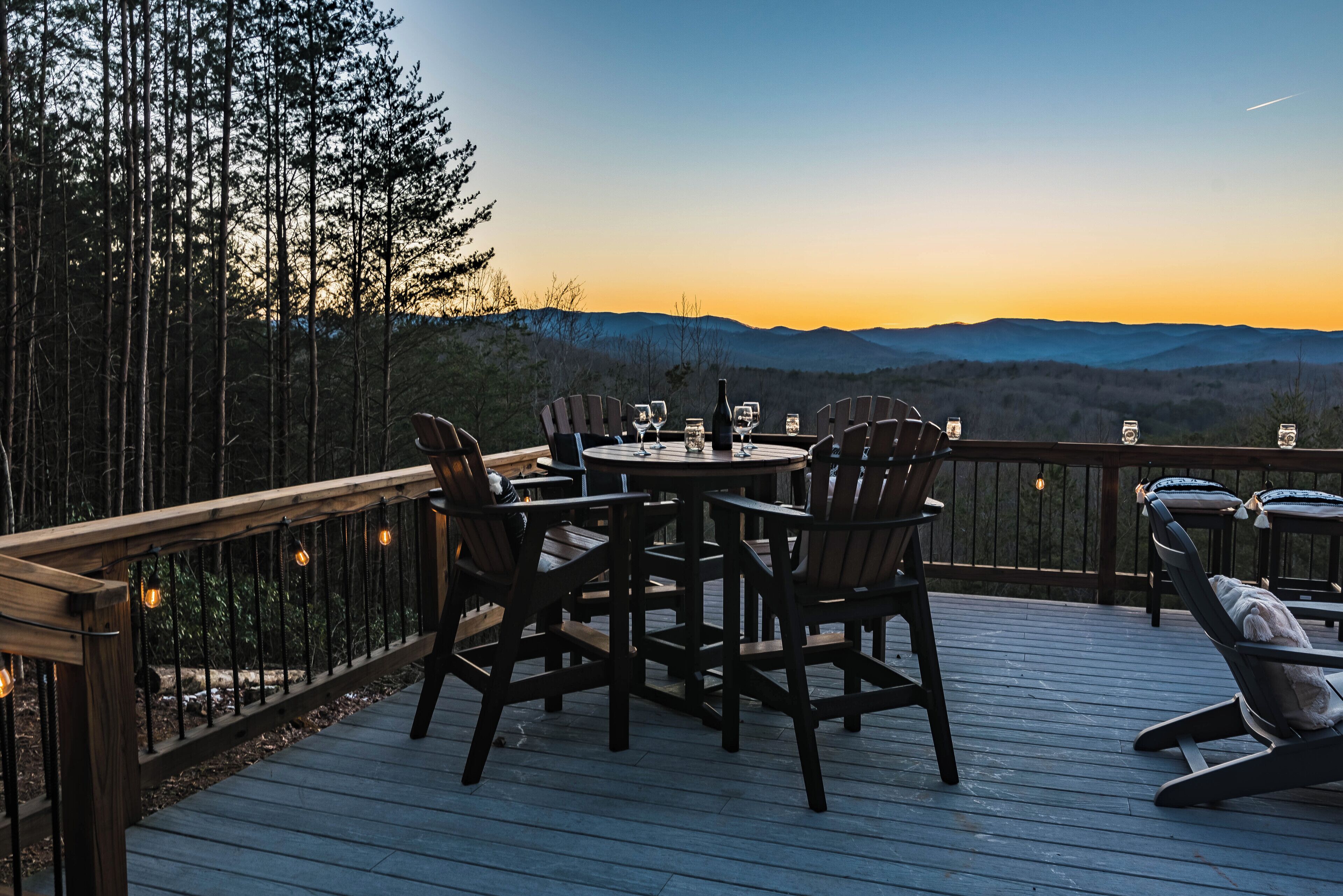 A deck with a round table and chairs, overlooking a mountainous landscape at sunset.