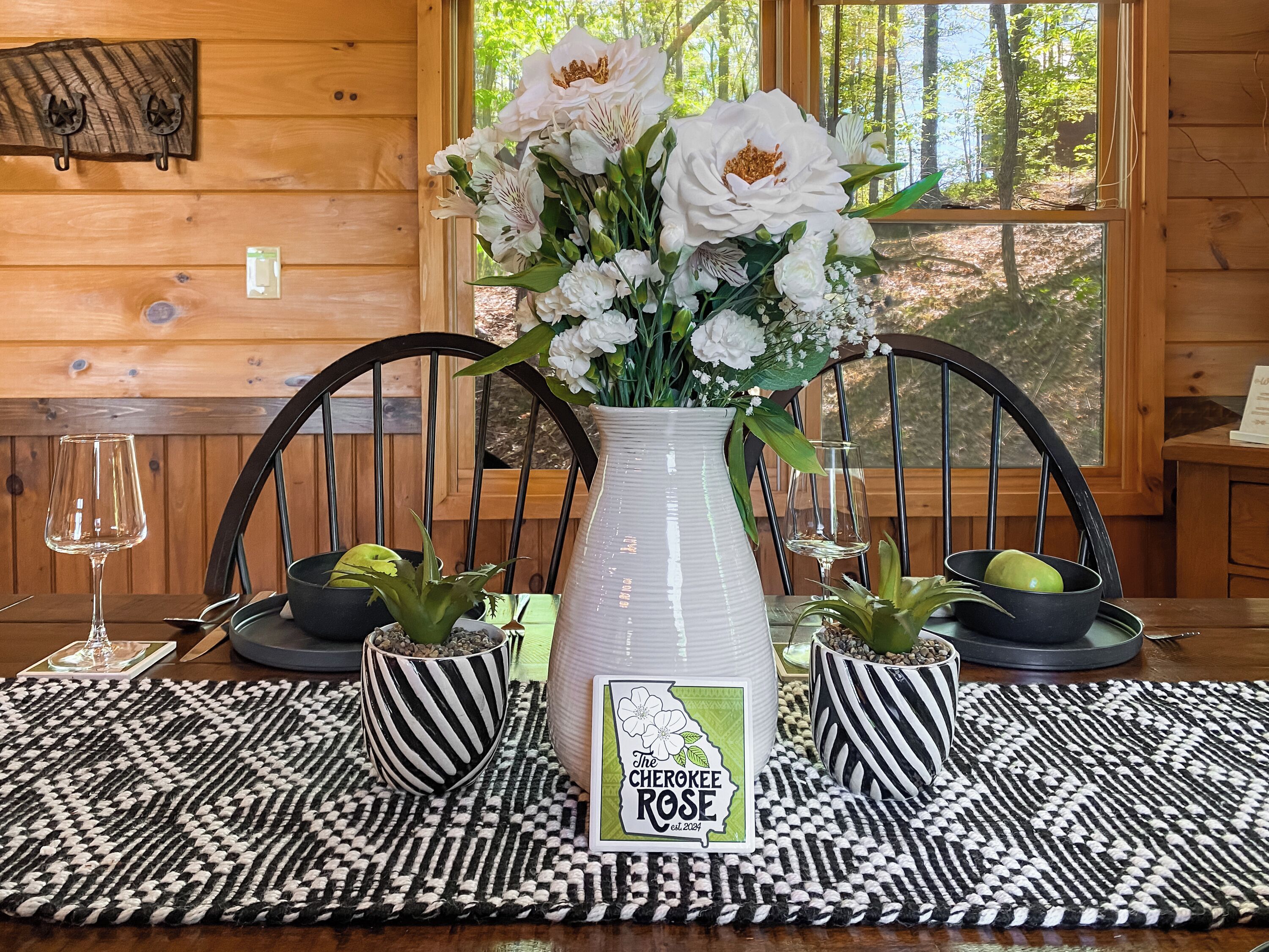 A wooden interior with a dining table set for two, featuring a vase of white flowers, black and white striped bowls, and a placemat with a logo.