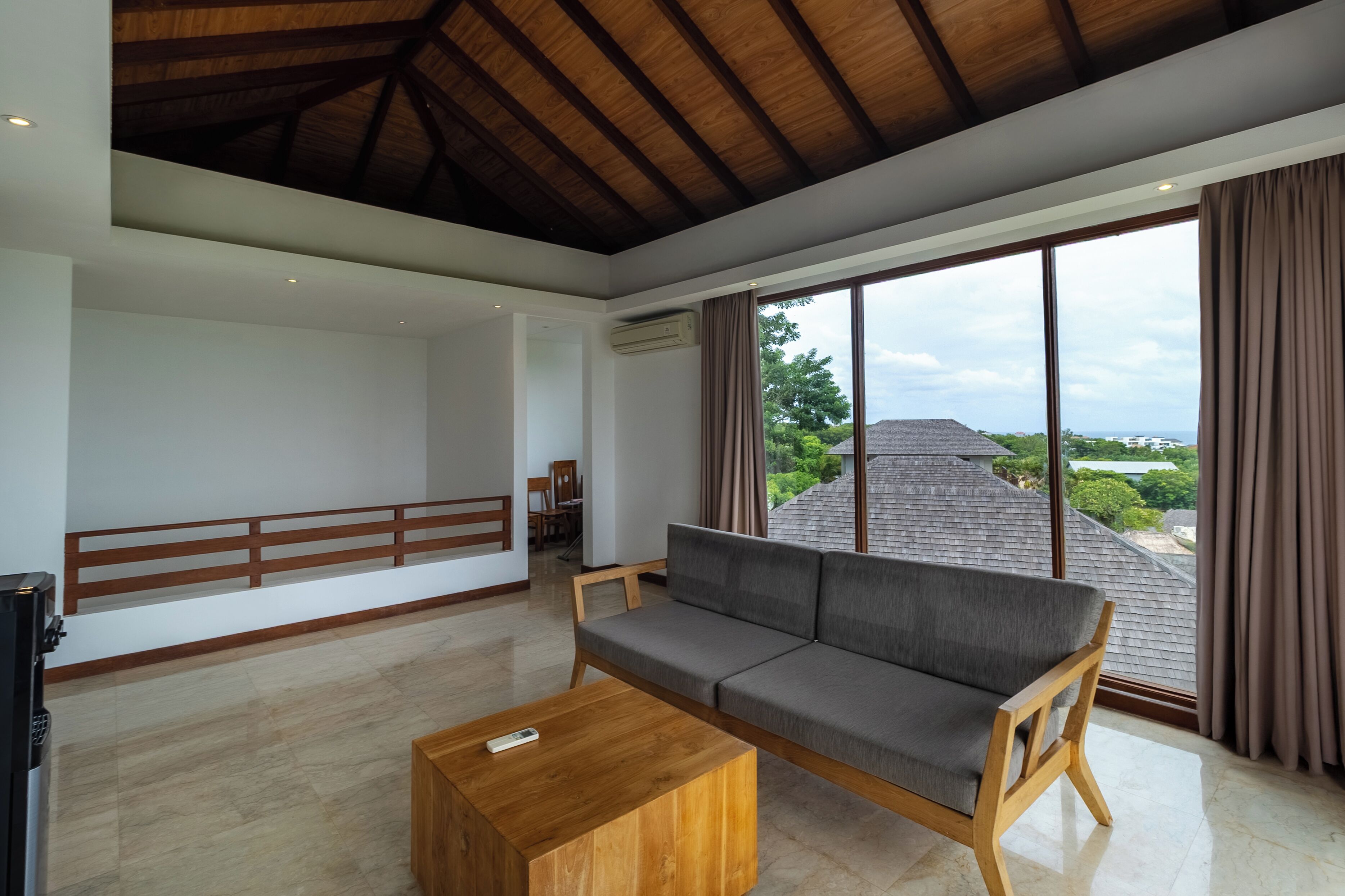 A spacious living area with a wooden ceiling, a grey sofa, a wooden coffee table, and large windows offering a view of the roof and greenery.