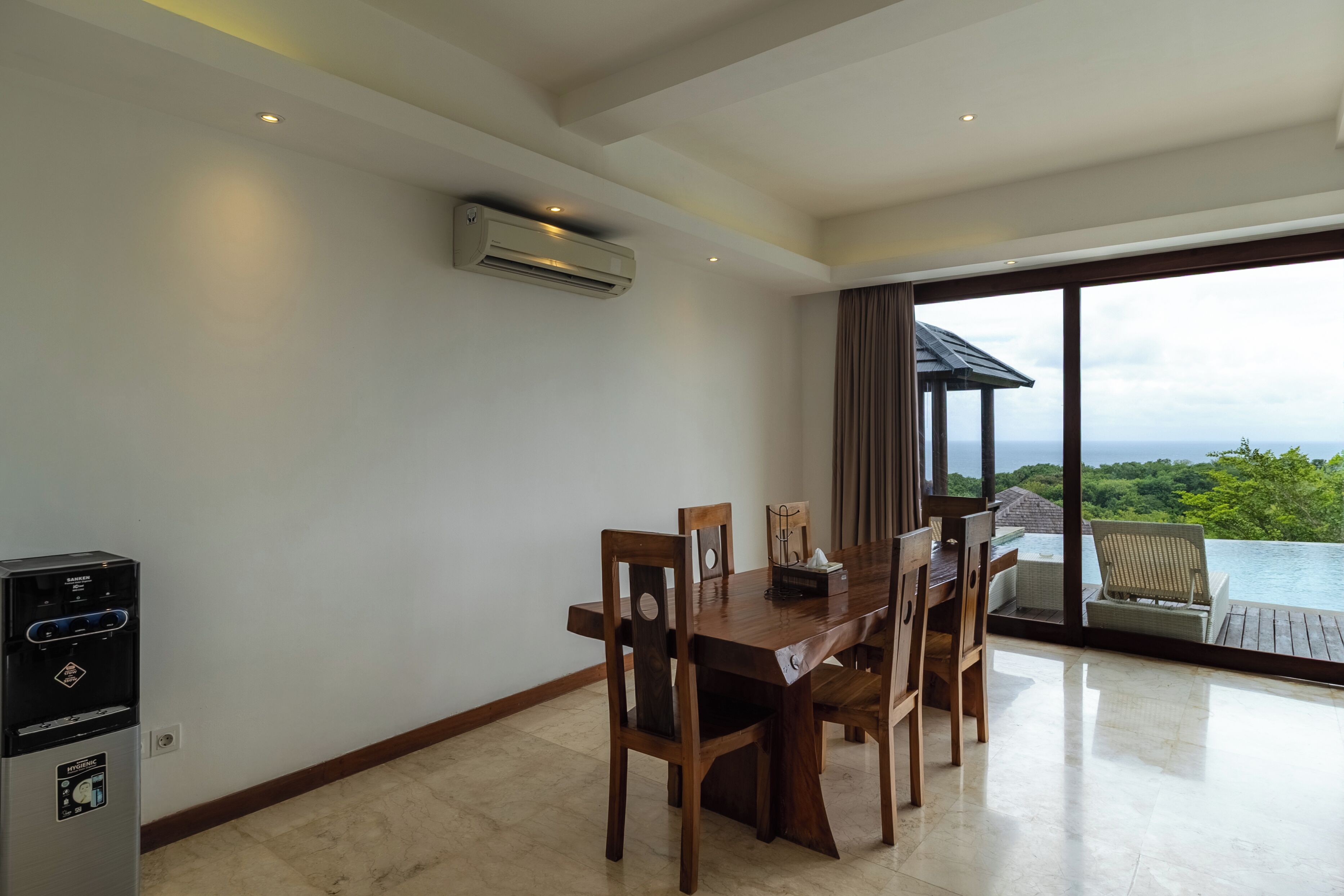 A dining area with a wooden table and chairs, an air conditioning unit, and a view of a lush landscape through large windows.