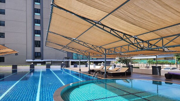 A swimming pool with a sunshade structure, lounge chairs, and a modern building in the background. - One Residence (Khlong Luang)