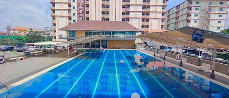 A swimming pool with a clear blue water surface, surrounded by a covered area with a roof and a building with multiple balconies in the background.