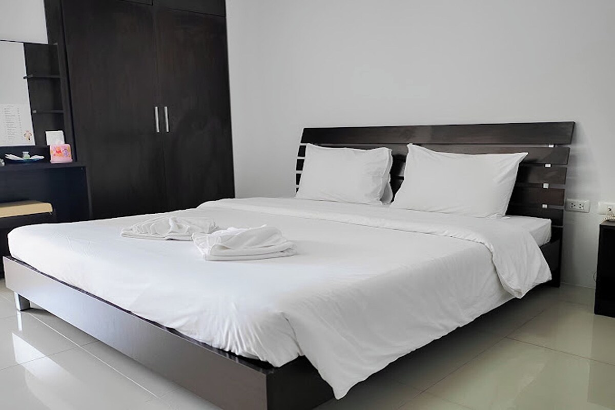 A modern bedroom with a large bed, dark headboard, white bedding, and a nightstand with a lamp.