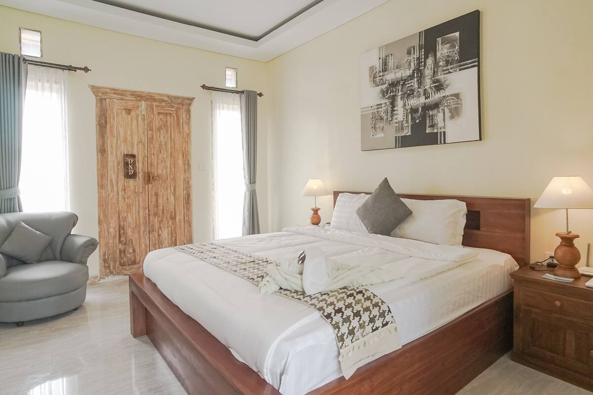 A bedroom with a large bed, two bedside tables with lamps, a grey armchair, a wooden wardrobe, and a wall-mounted abstract art piece.