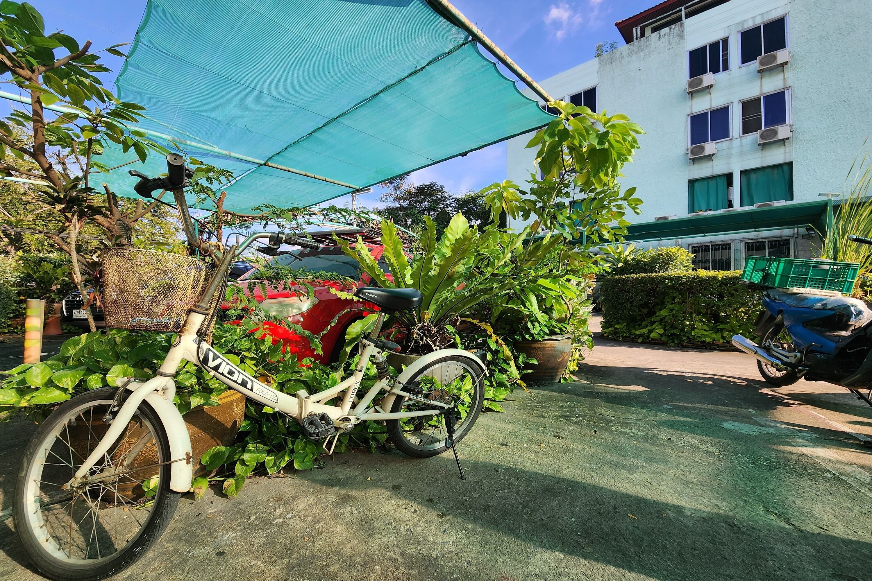 Una bicicleta aparcada bajo una estructura de sombra, rodeada de vegetación y un edificio al fondo.