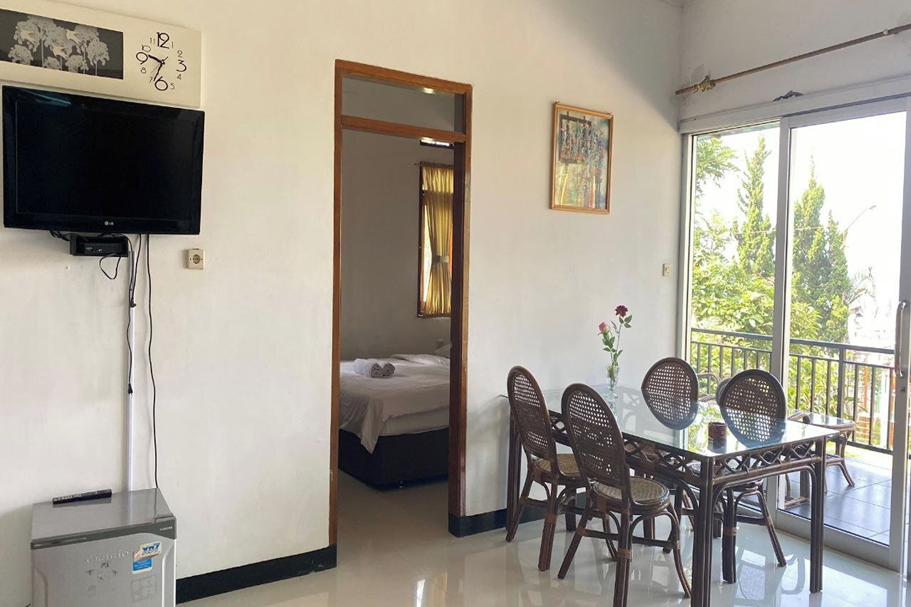 A hotel room with a bed, a dining table with chairs, a TV mounted on the wall, and a balcony with a view of trees.