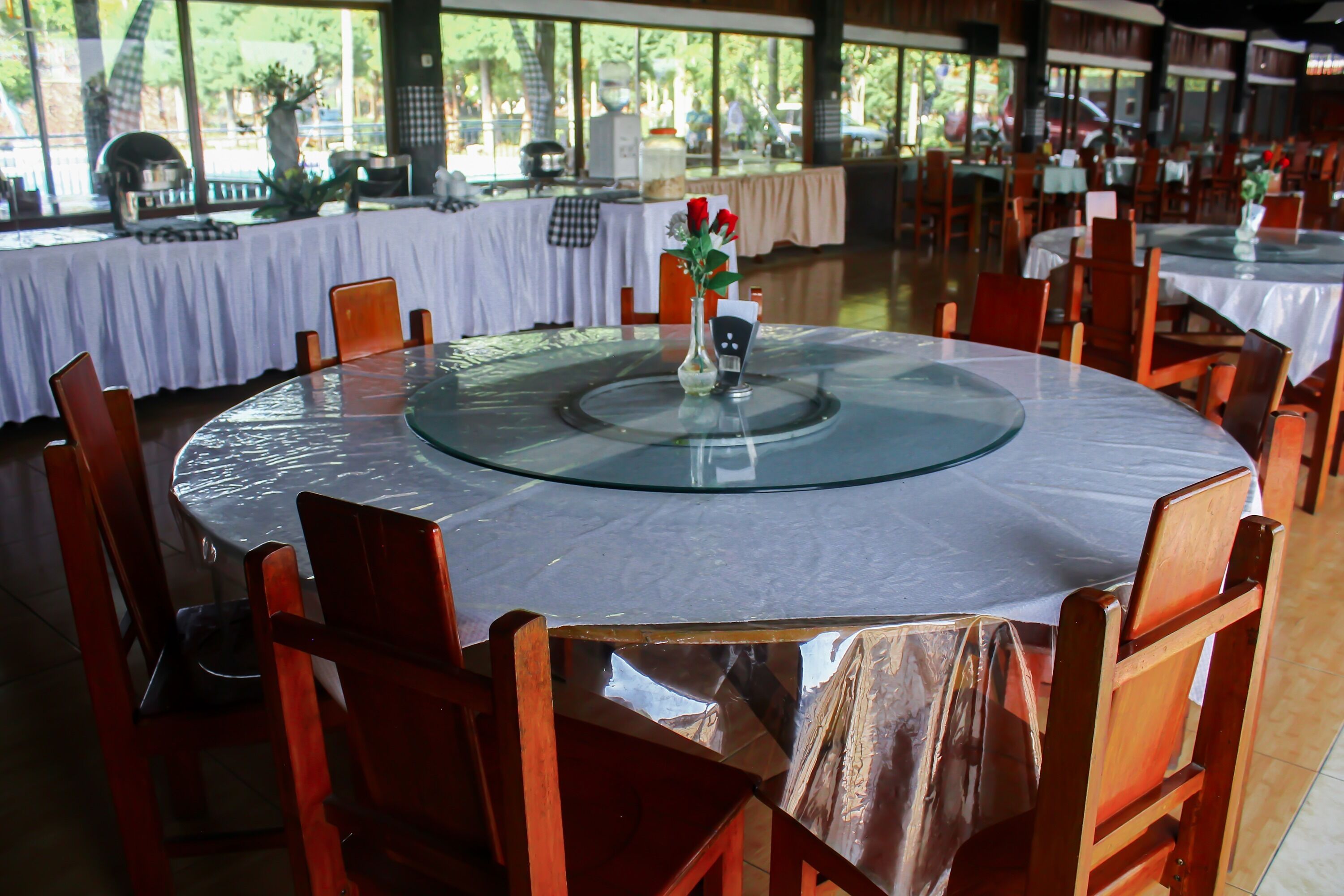 A restaurant with round tables covered in white tablecloths, wooden chairs, and a vase with flowers on one table.