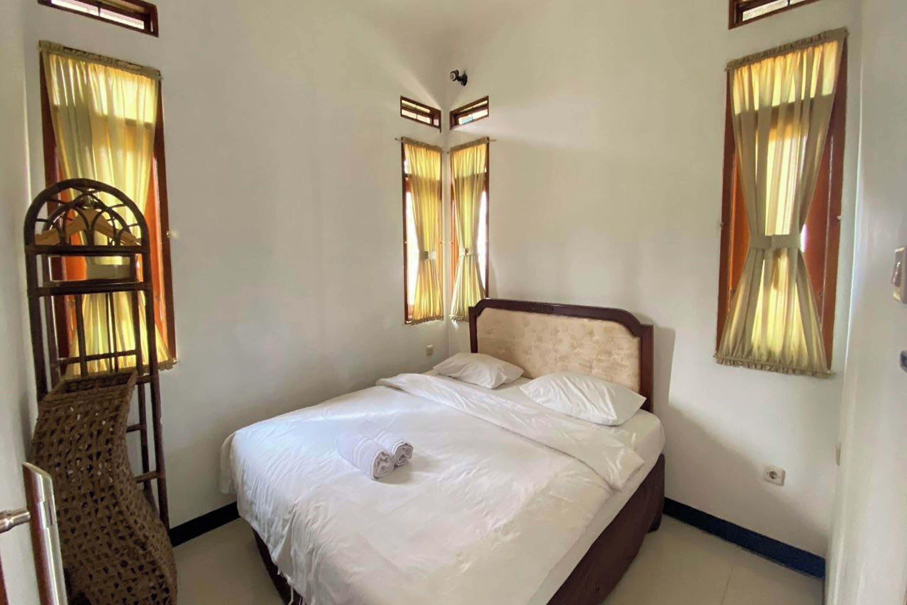 A neatly made bed with white linens and a pair of towels on the bed. There are two windows with curtains, and a small decorative item is visible on the wall.