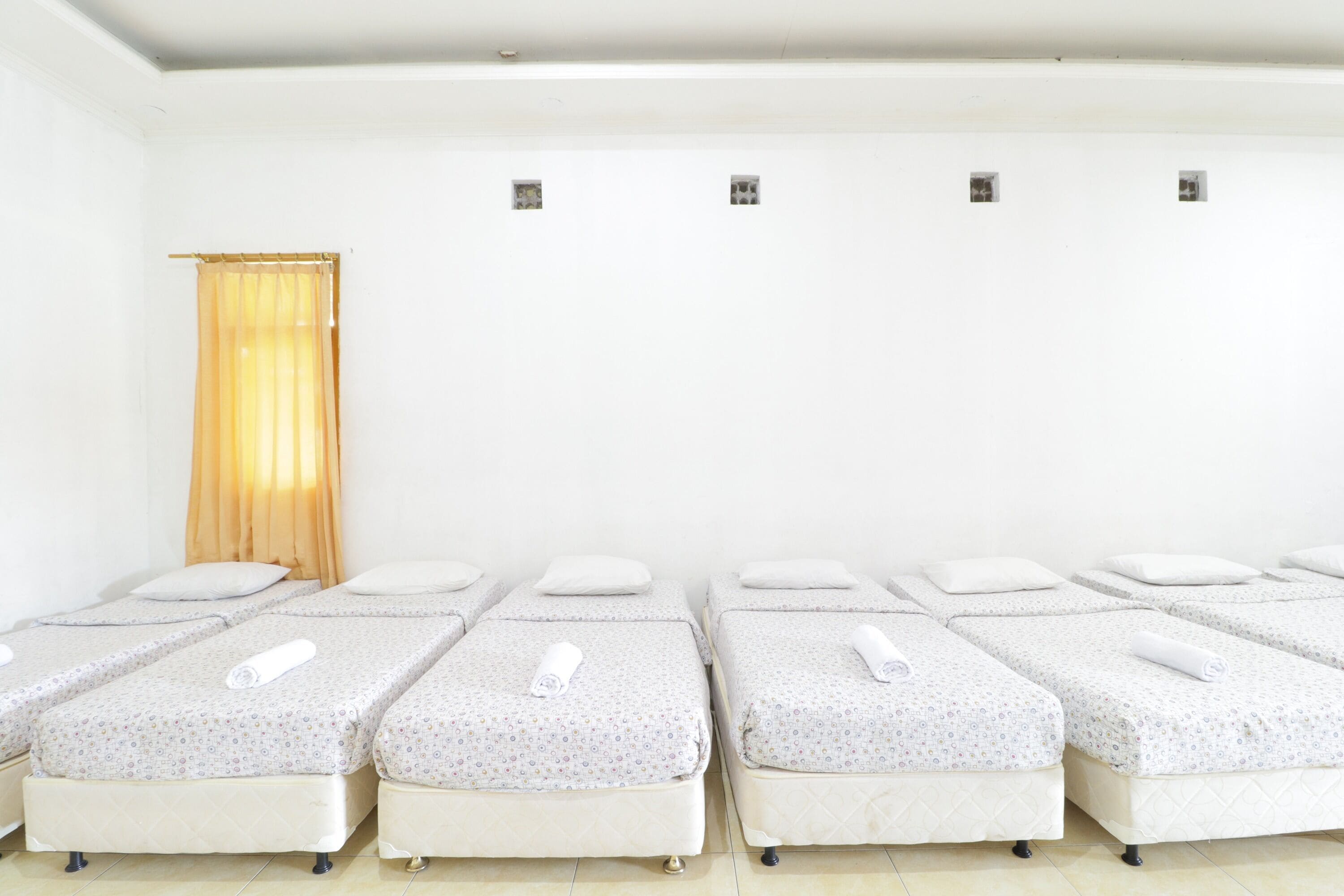 A row of four single beds with white linens and a patterned duvet cover, arranged in a room with white walls and a wooden floor.