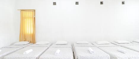 Cuatro camas individuales con sábanas blancas y una colcha de diseño, dispuestas en una habitación con paredes blancas y suelo de madera.