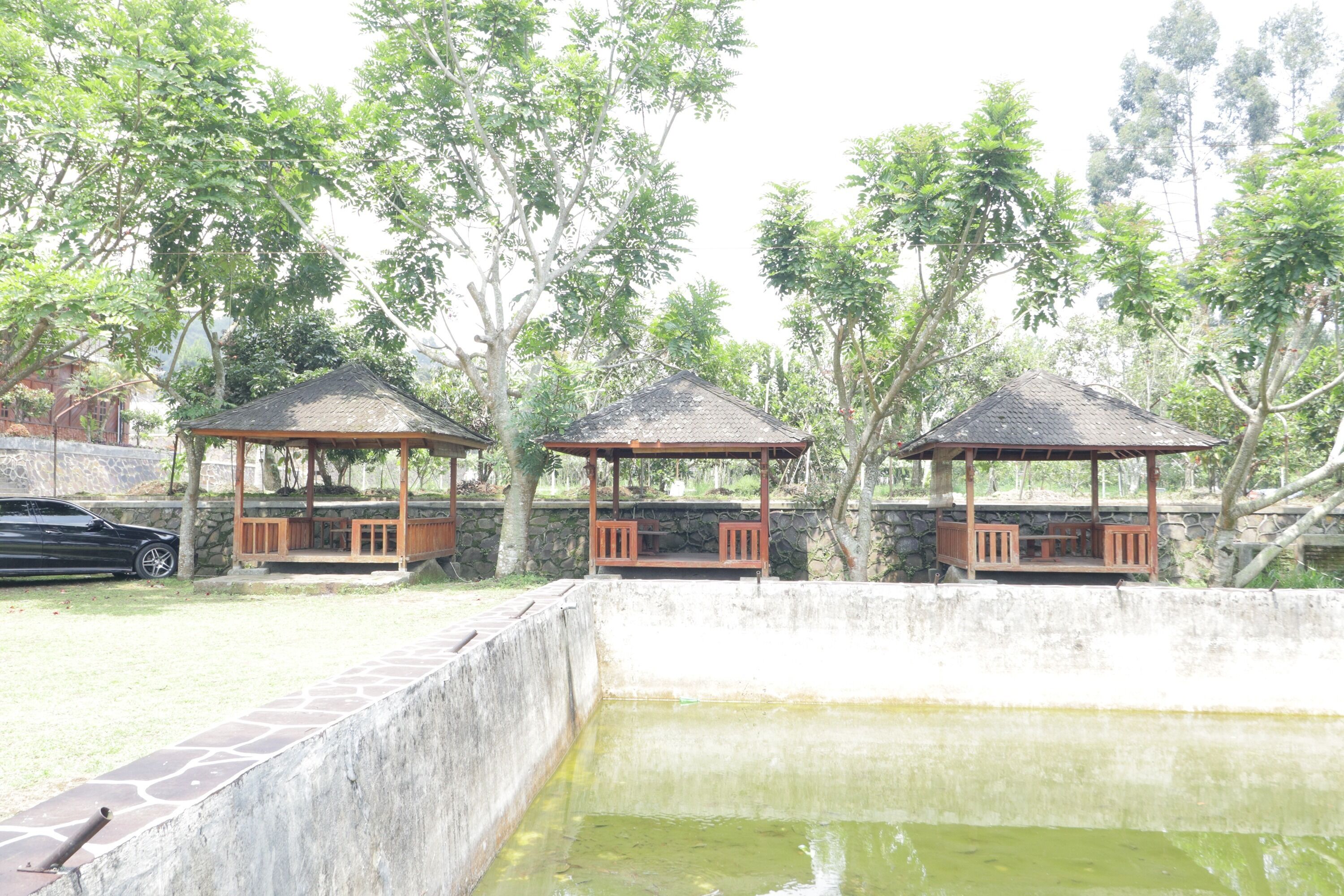 A gazebo structure with a stone base and a tiled roof, surrounded by trees and a concrete wall.