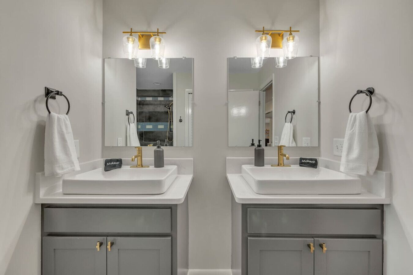 A mirrored bathroom with two sinks, each with a gold faucet and a makeup station.