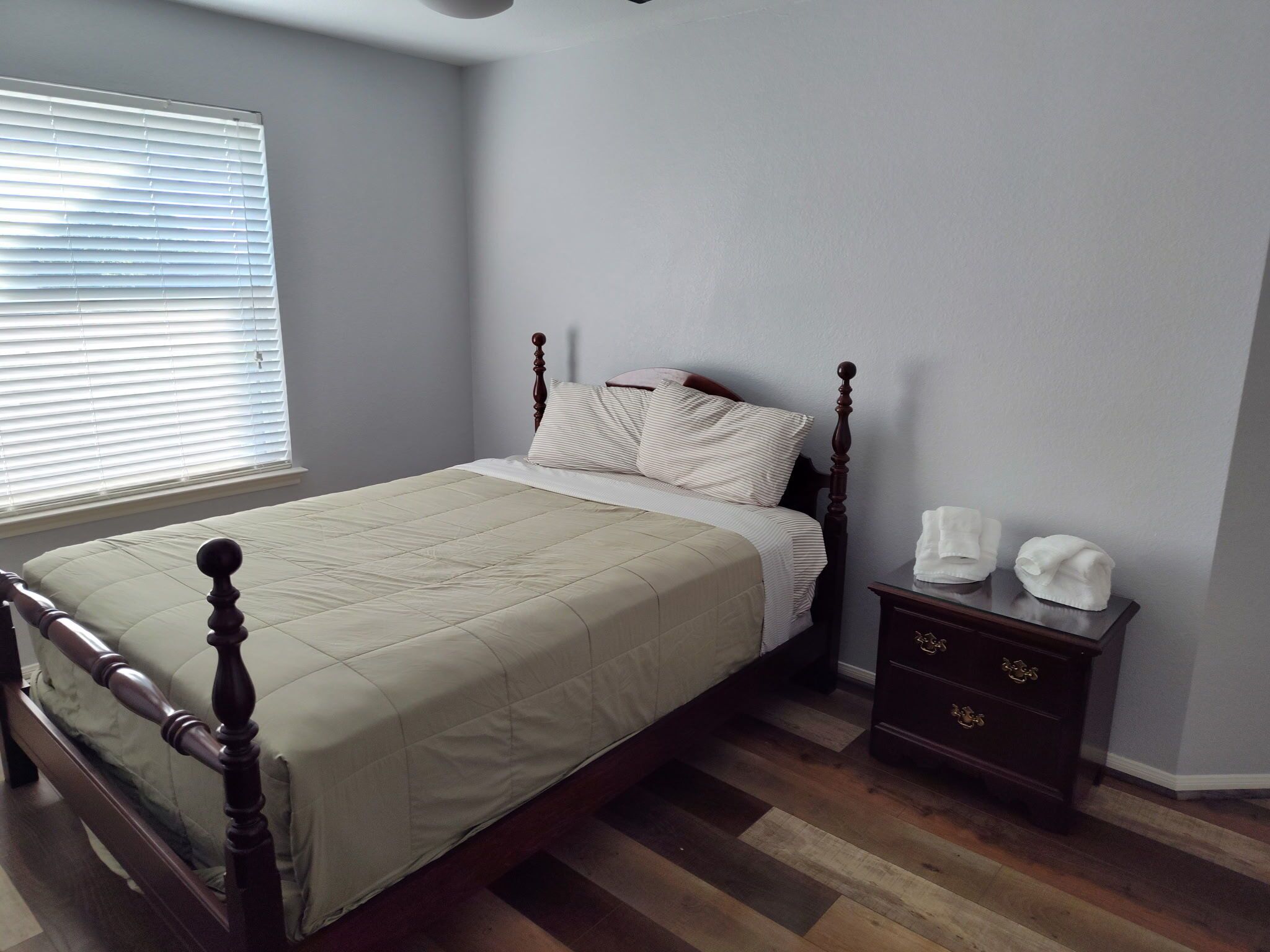 A bedroom with a bed, a nightstand, a window with blinds, and a wooden floor.