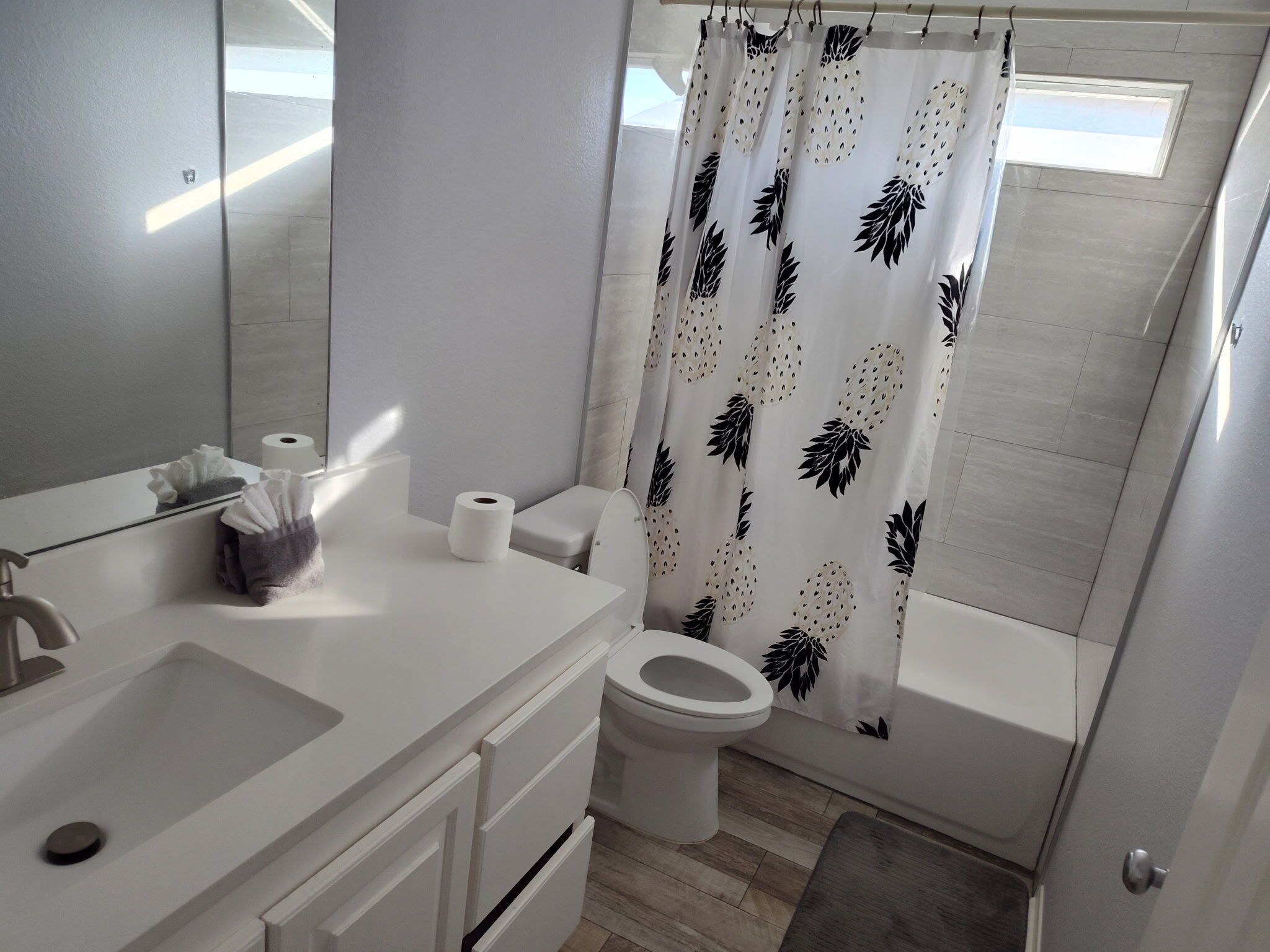 Ein modernes Badezimmer mit einem weißen Waschbecken, einer Toilette und einer Badewanne. Ein Duschvorhang mit Blättermuster, ein Handtuchhalter mit Handtuch und Taschentuchbox.