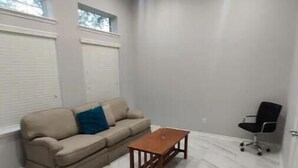 A room with a sofa, a wooden coffee table, a black office chair, and a window with blinds.