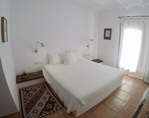 A bedroom with a large bed, a bedside table, a lamp, a small plant, a rug, and a window with curtains. - 5 Star Villa Can Perez – Hidden Gem in Ibiza’s North-East (Balearic Islands)