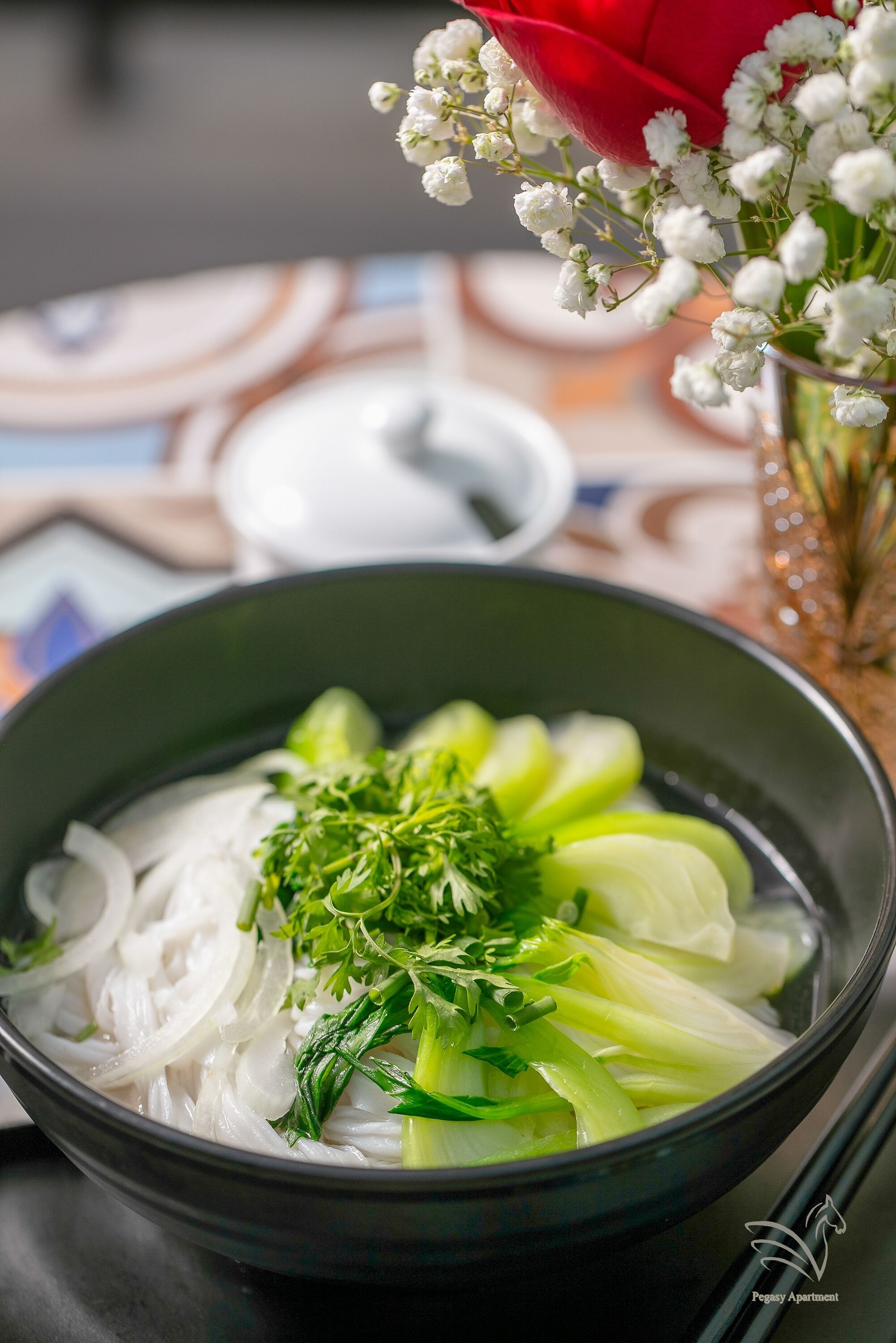 A bowl of clear soup with vegetables, a red rose, and white flowers in the background.