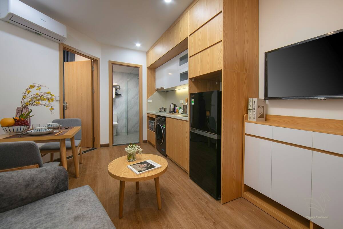 Une cuisine compacte avec des meubles en bois, un four encastré et un réfrigérateur noir. Une table à manger avec un vase de fleurs et un magazine se trouve au premier plan.