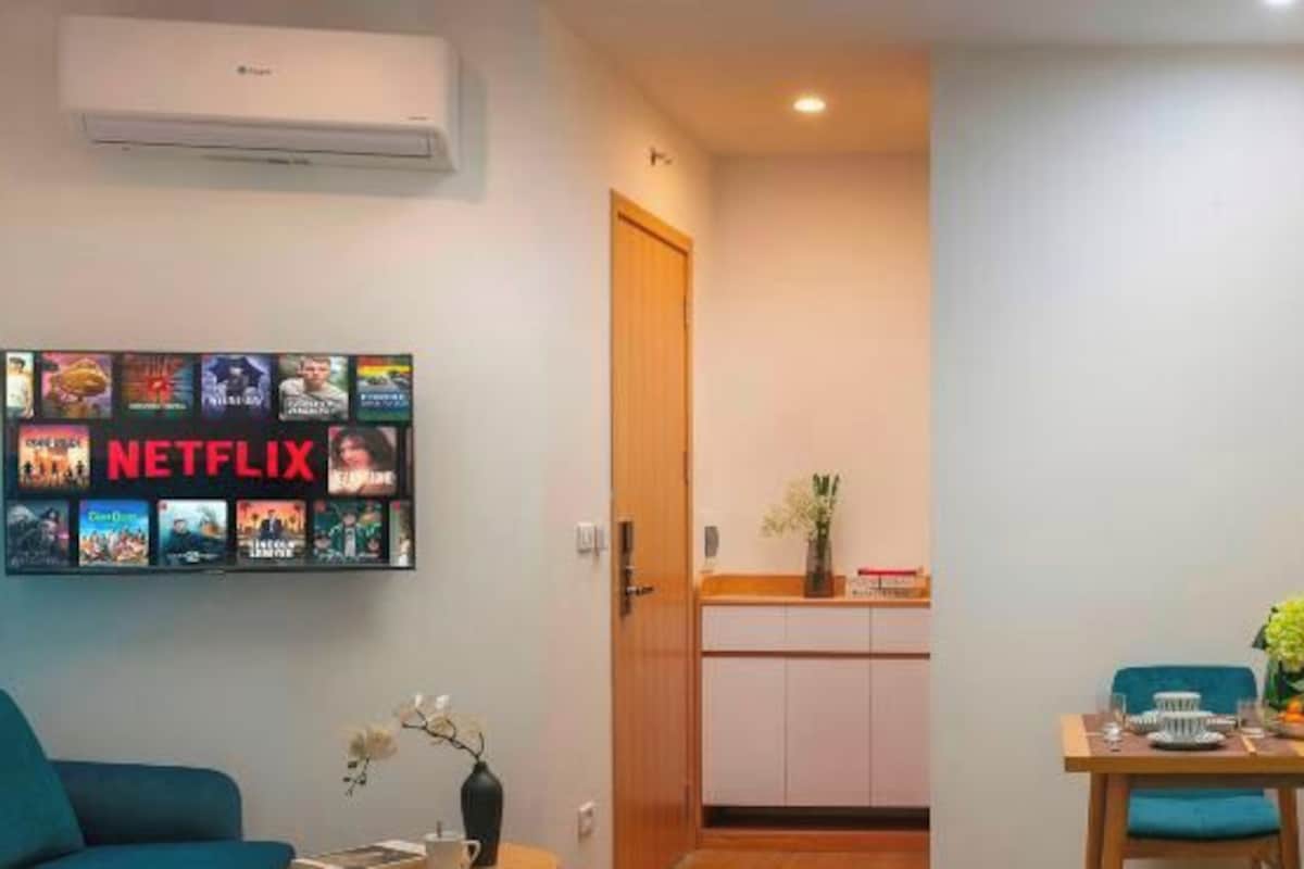 A living room with a wall-mounted TV displaying various Netflix shows, a sofa, and a small table with a vase and flowers.