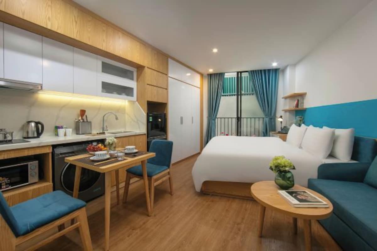 A compact hotel room with a kitchenette, a bed, a seating area, and a view of the outside.