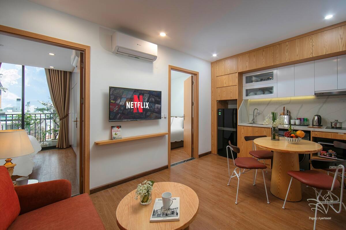 Une chambre d’hôtel moderne comprenant un coin salon, une table à manger avec quatre chaises, une cuisine avec des meubles en bois et un téléviseur fixé au mur affichant le logo de Netflix.