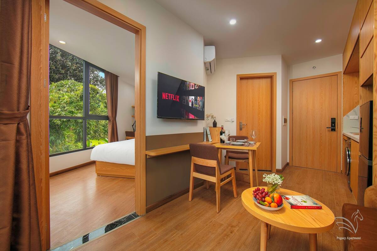 Une chambre d’hôtel moderne équipée d’un lit, d’un téléviseur affichant l’application « NETFLIX », d’un coin repas avec une table et des chaises, et offrant une vue sur la verdure par la fenêtre.