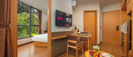 Habitación de hotel moderna con cama, televisor mostrando 'NETFLIX', zona de comedor con mesa y sillas, y vista a zonas verdes por la ventana.