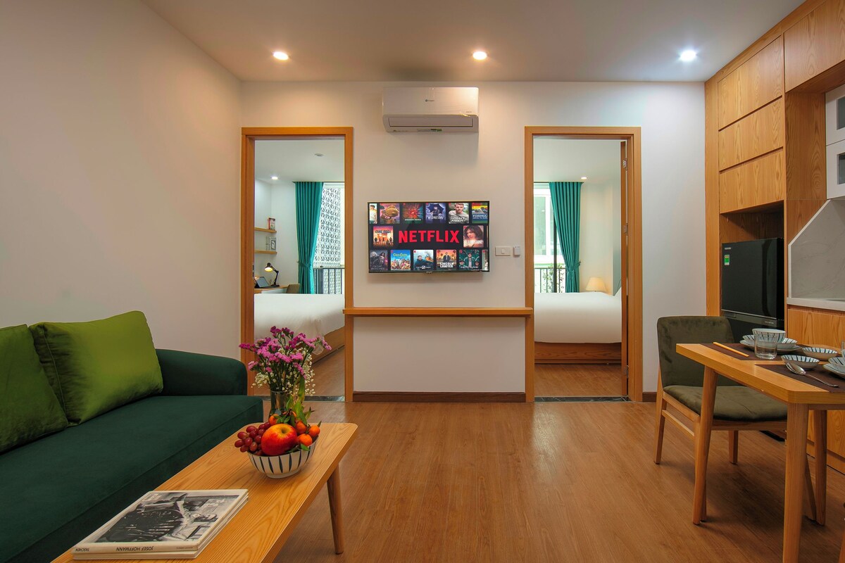 A modern living room with a wooden coffee table, a sofa, a dining table with chairs, a kitchen area, and a wall-mounted TV displaying the Netflix logo.