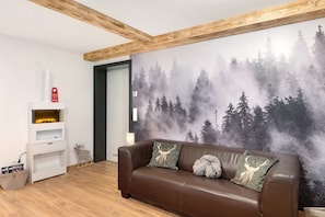 A room with a brown leather sofa, a white shelving unit, a fireplace, and a forest-themed wall mural.