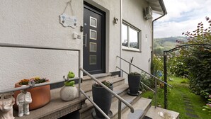 Fachada de una casa con una puerta oscura, escalones con barandilla de metal, plantas en macetas y vistas a un paisaje verde.