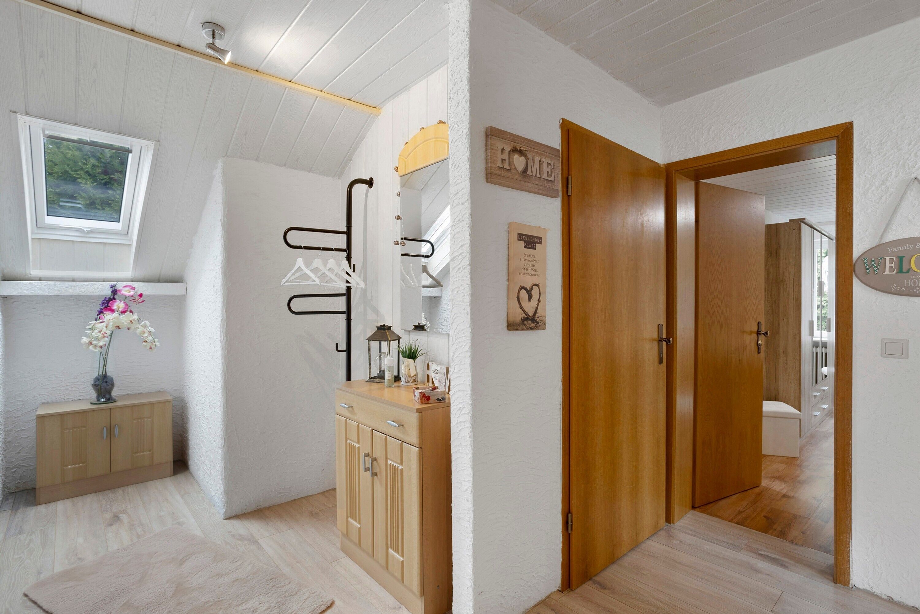A hallway with wooden doors, a coat rack, a small cabinet with a vase, and a window with a view of greenery.