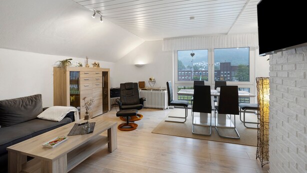 Una sala de estar moderna con sofá, mesa de centro, comedor y vista a la ciudad a través de amplias ventanas.