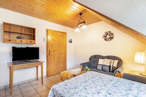 Ein Zimmer mit einem Bett, einem Fernseher auf einem Holztischchen, einer Holztür, einem Wandregal und einer Deckenleuchte.