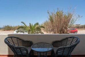Een balkon met rieten stoelen en een glazen tafel, met uitzicht op een parkeerplaats met een rode auto en begroeiing.