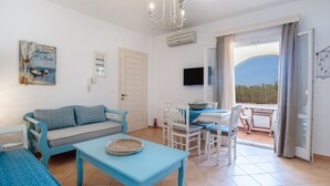 Een kamer met een bank, een eettafel met stoelen, een aan de muur bevestigde televisie en een balkon met gordijnen.
