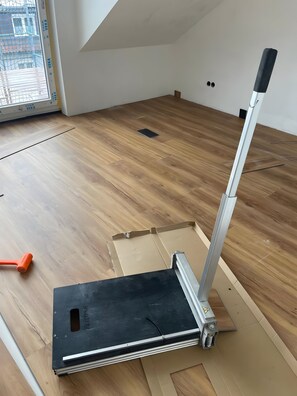 Une pièce avec un plancher en bois, une fenêtre avec des stores et un meuble dont le dessus est noir.